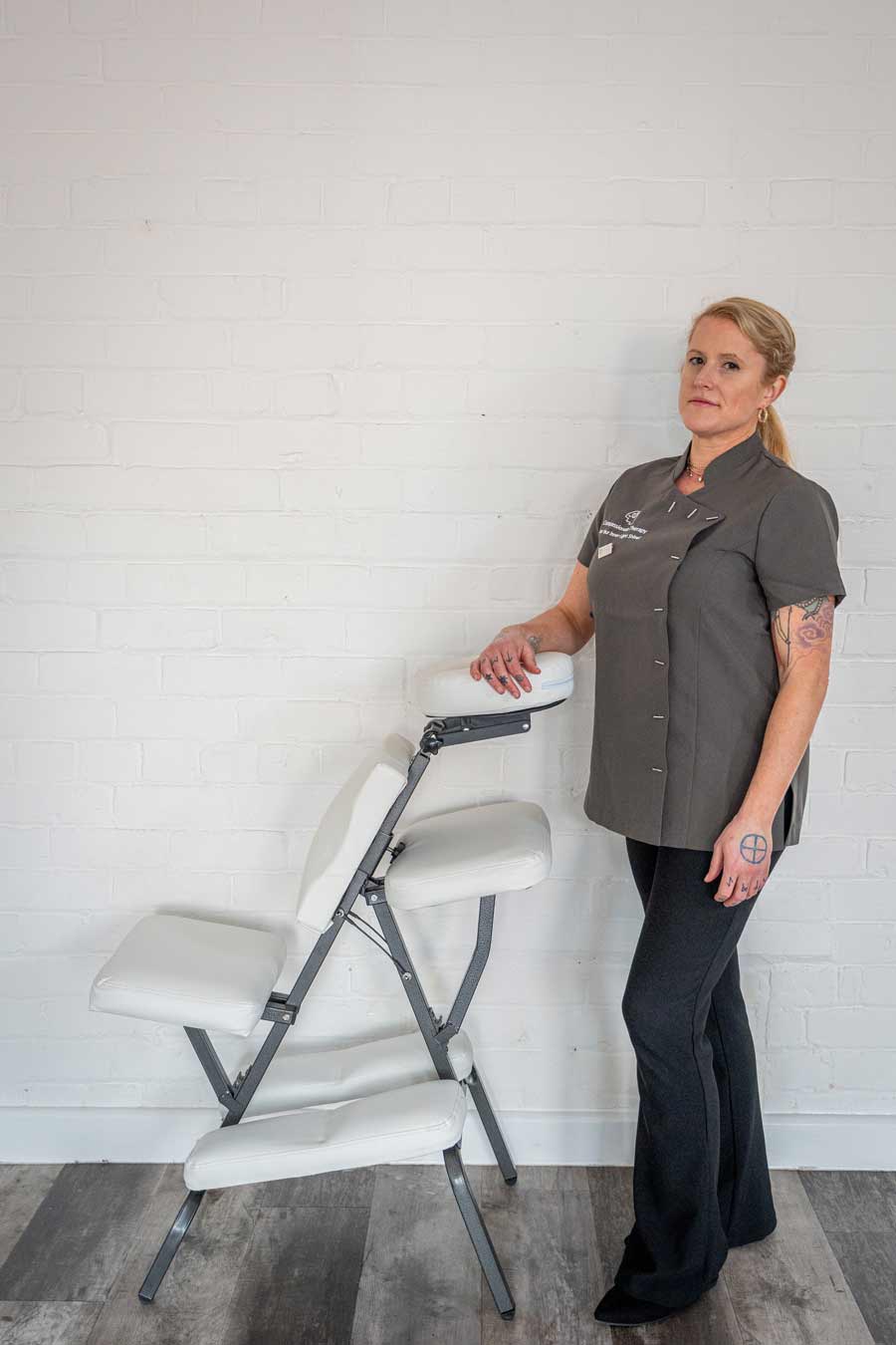 A woman in a gray uniform stands indoors beside a white massage chair, suggesting a setting for coaching or training, with one hand resting on it against a white brick wall background.