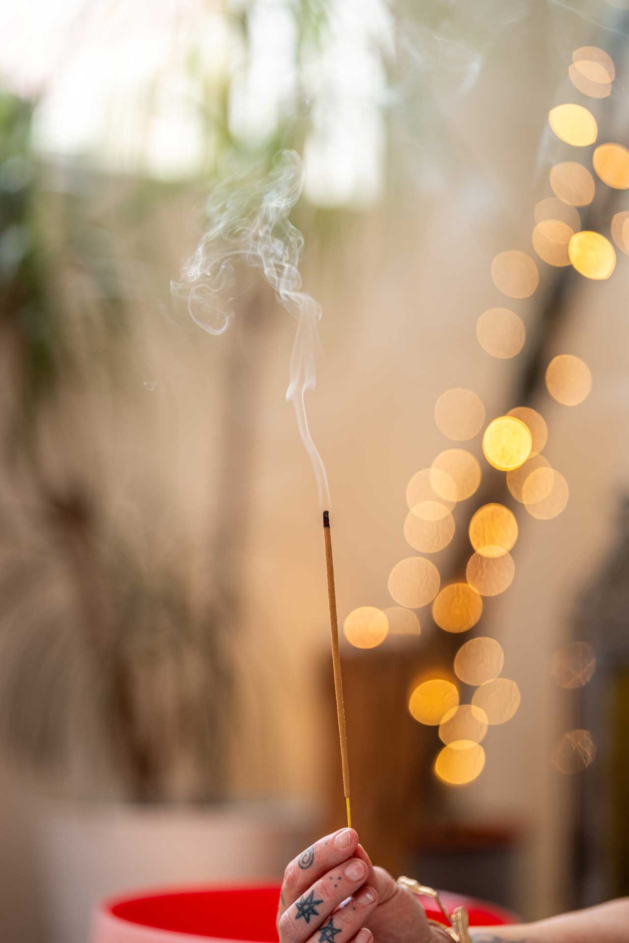 A hand with tattoos holds a burning incense stick, releasing smoke—a serene moment of mindfulness. The background is softly blurred with warm, circular lights and hints of greenery.